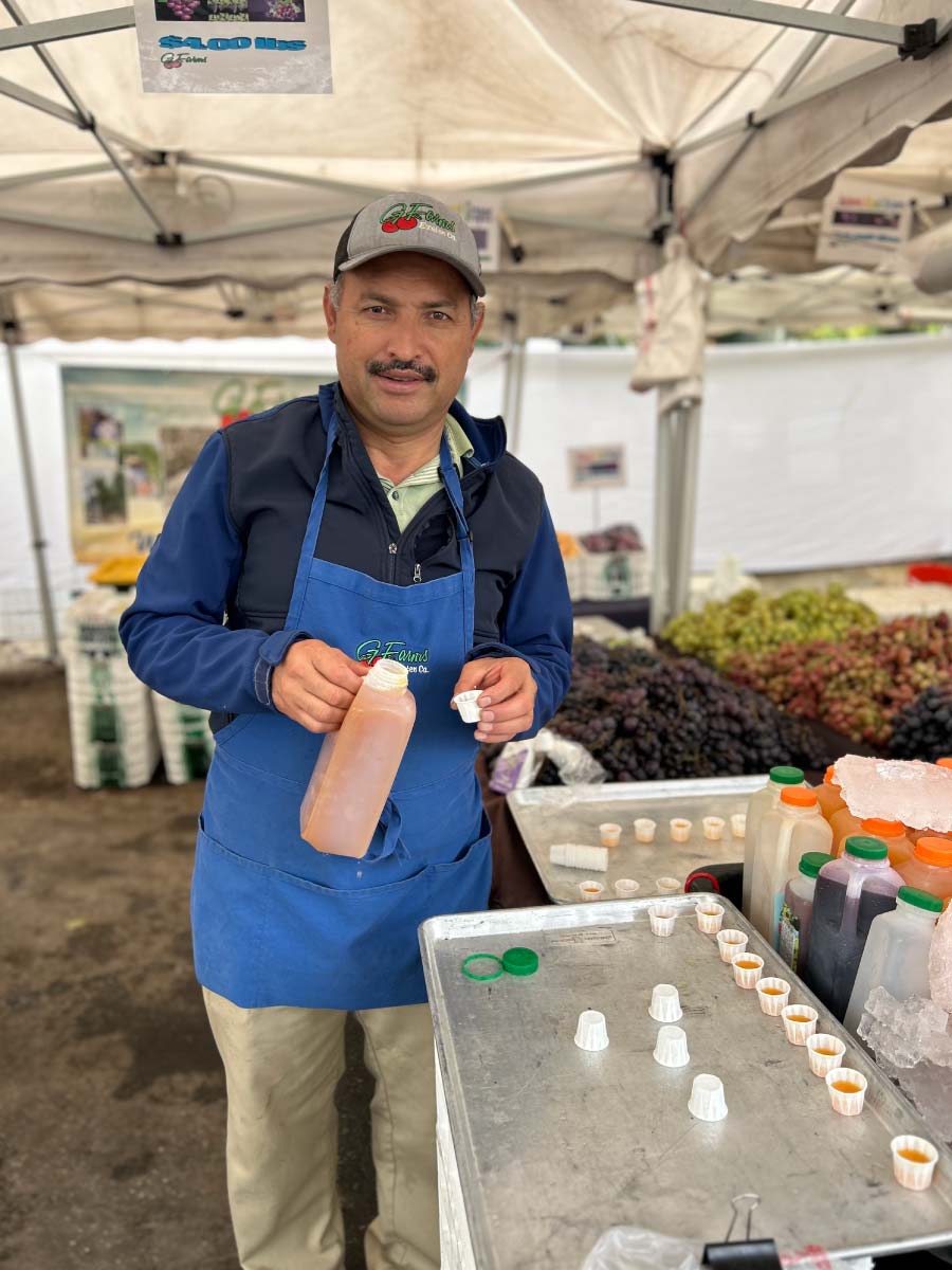 A man wearing a blue G Farms apron and baseball cap stands at a market stall, pouring a light-colored juice from a large plastic jug into small sample cups. In the background, crates of purple and green grapes are displayed under a white canopy.
